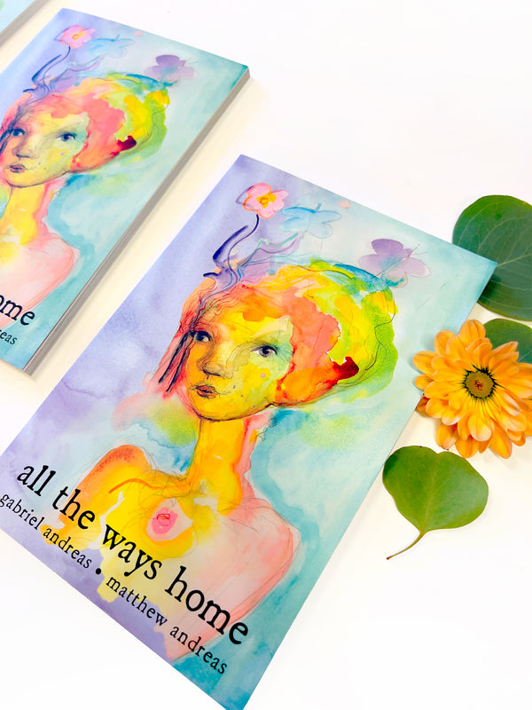 Two copies of the book 'all the ways home' by gabriel andreaes & matthew andreaes, featuring a colorful watercolor illustration of a woman with vibrant hair, styled on a white surface with green leaves and an orange flower.