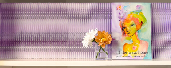 A book titled 'all the ways home' by gabriel andreas - matthew andreas leaning against a purple striped wall on a white shelf, next to a small vase with white and orange flowers.