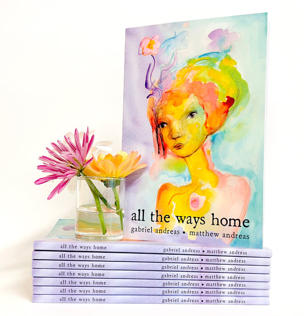 A book titled 'all the ways home' by gabriel andreas and matthew andreas is shown standing upright next to a stack of identical copies, with a small glass vase of pink and yellow flowers in front. The cover features a colorful watercolor portrait of a woman with vibrant orange, yellow, green, and purple tones on a light background.