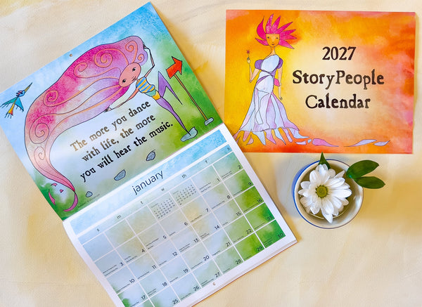 An illustrated 2027 StoryPeople Calendar shown open on a light wood surface, with the cover page displaying a pink figure and  the calendar grid for January visible. A small white flower in a blue dish sits beside it.